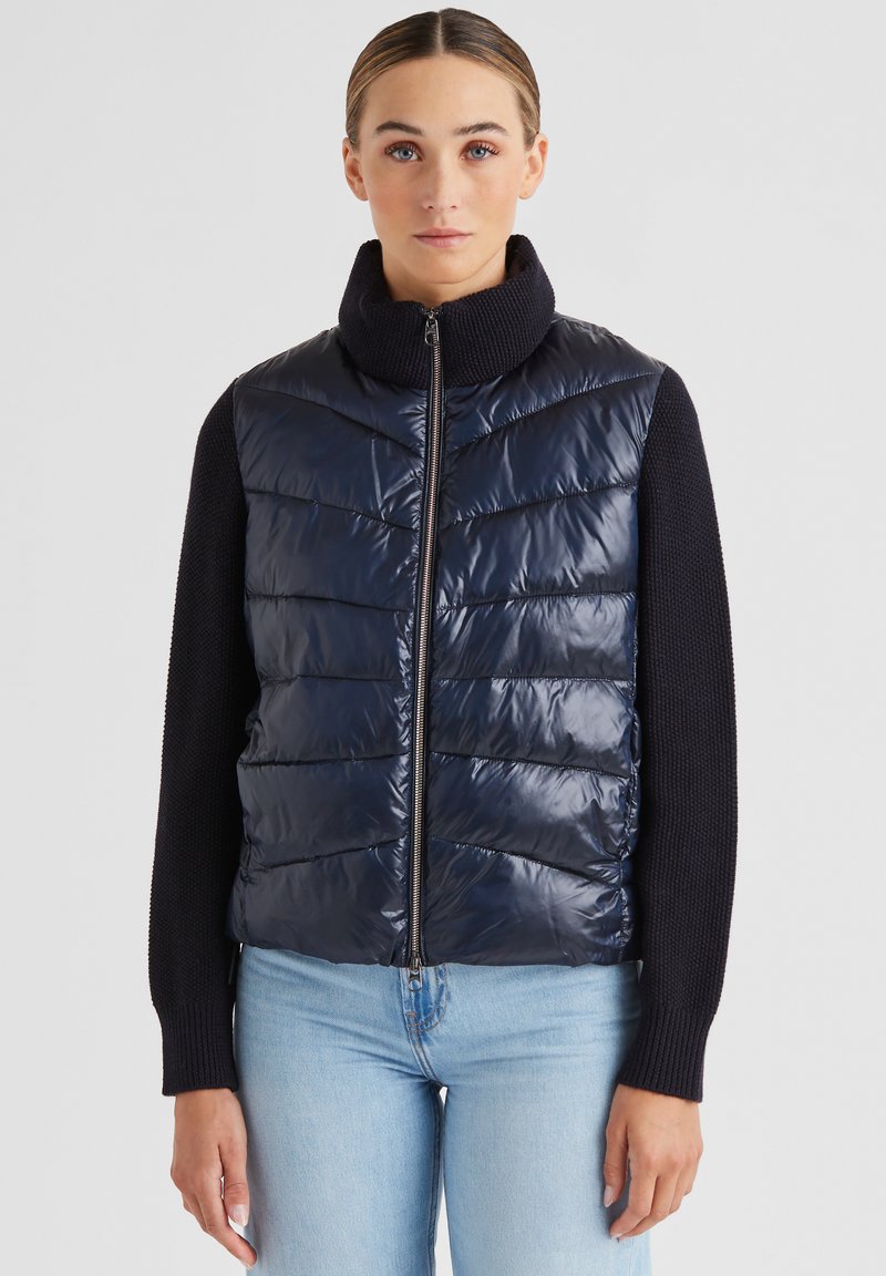 Navyblauer Steppwesten mit hohem Kragen, Reißverschluss und horizontalem Steppmuster; gerippte schwarze Ärmel.