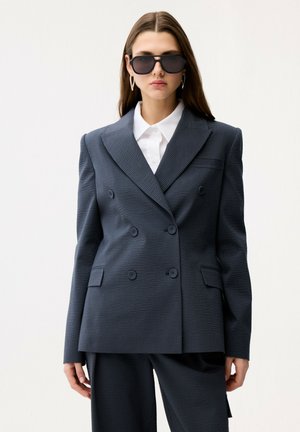 Kvinde iført en oversized tekstureret mørkegrå dobbeltknappet blazer, matchende bukser, hvid skjorte med krave, store sorte solbriller og sølvfarvede hoop-øreringe.