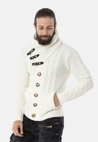 Weißer Strickpullover mit hohem Kragen, ausgestattet mit schwarzen Knotenverschlüssen und großen braunen Knöpfen, die strukturierte Muster zeigen.