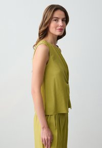Top senza maniche di colore verde oliva con design drappeggiato e texture morbida, abbinato a pantaloni coordinati, mostrato da un lato su uno sfondo neutro.