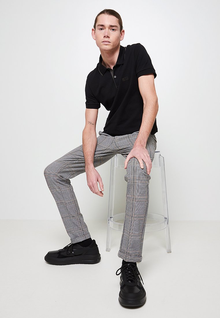 T-shirt polo noir, pantalon slim-fit à carreaux gris, baskets noires, assis sur une chaise transparente avec un arrière-plan neutre. Design simple et coupe moderne.