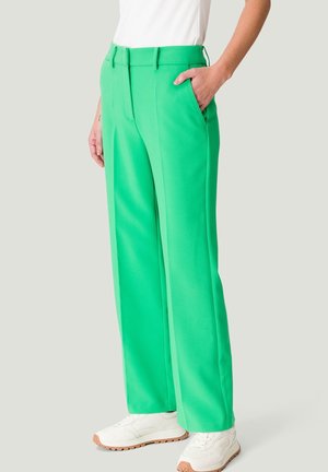 Woman wearing bright green wide-leg trousers, white sneakers, and a white top, standing with one hand in pocket against a plain background.