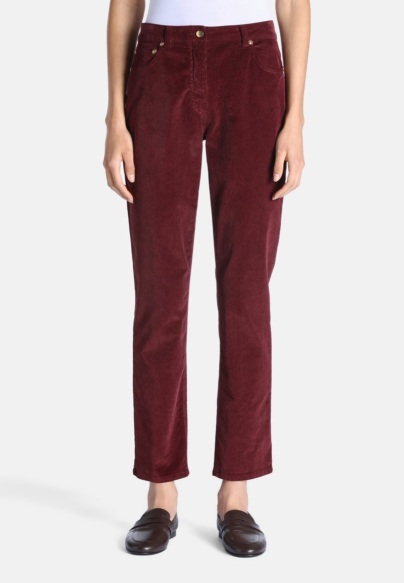 Personne portant un pantalon droit en velours côtelé bordeaux, un haut blanc et des chaussures marron foncé sans lacets, debout sur un fond blanc uni.