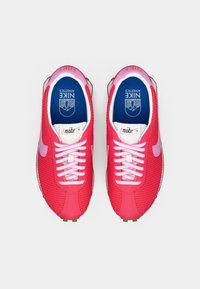 Um par de sapatilhas de corrida Nike cor-de-rosa brilhante com swooshes roxos, atacadores brancos e palmilhas azuis rotuladas "Nike Athletics" vistas de cima.