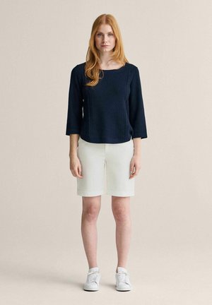 Kvinde med rødt hår iført en marineblå striktrøje, hvide knælange shorts og hvide sneakers, stående mod en ensartet lys baggrund.