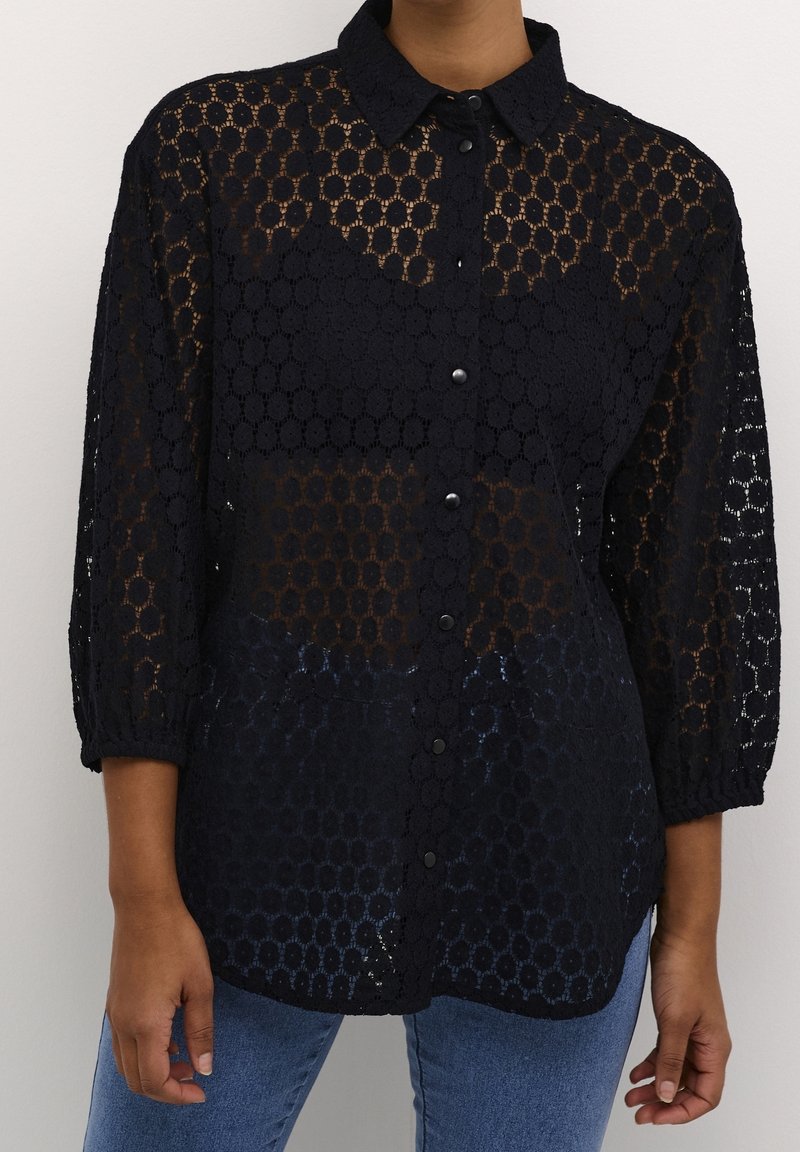 Chemise noire boutonnée en tissu à motifs de dentelle circulaire, portée avec un jean bleu, présentant des manches trois-quarts et un col à revers.