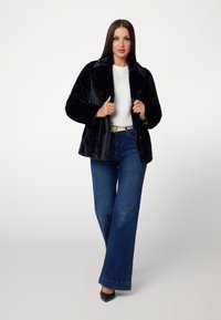 Giacca nera in finta pelliccia con colletto, chiusura senza bottoni, abbinata a un maglione bianco texturizzato e jeans a gamba larga blu. Silhouette definita.