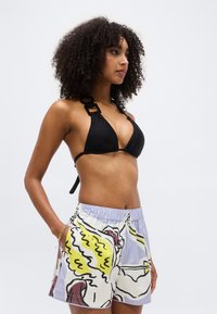 Mujer con cabello rizado, vistiendo un top de bikini negro y pantalones cortos con estampado colorido, de pie con una mano en el bolsillo, mirando hacia la derecha.