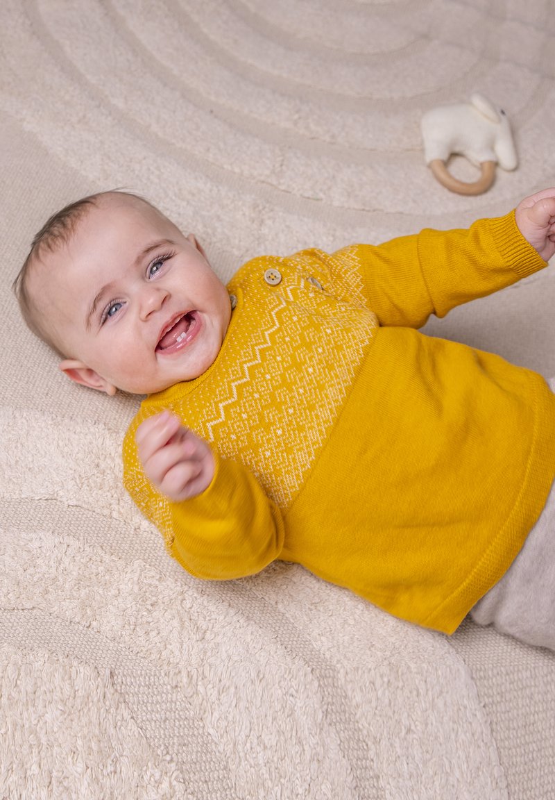 Maglione giallo lavorato a maglia con motivo geometrico bianco, presenta un bottone vicino al colletto, indossato da un neonato sdraiato su una superficie testurizzata di colore chiaro.