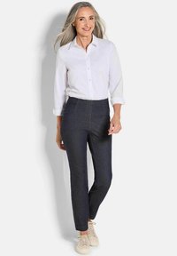 Lachende vrouw met lang grijs haar, gekleed in een witte knoopshirt, donkere slim fit broek en beige sneakers, staande voor een effen achtergrond.