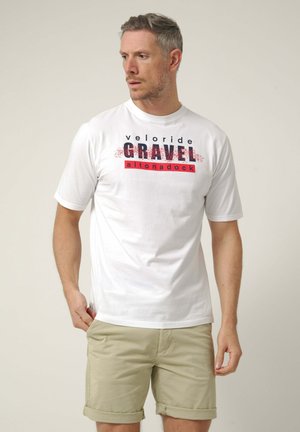 Hombre con el pelo corto que lleva una camiseta blanca "GRAVEL altonadock" y pantalones cortos beige, mirando hacia su izquierda sobre un fondo liso.
