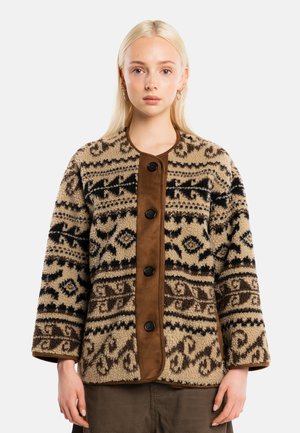 Mujer joven con pelo rubio liso que lleva una chaqueta de forro polar beige y marrón con botones y pantalones marrones, de pie frente a un fondo liso.
