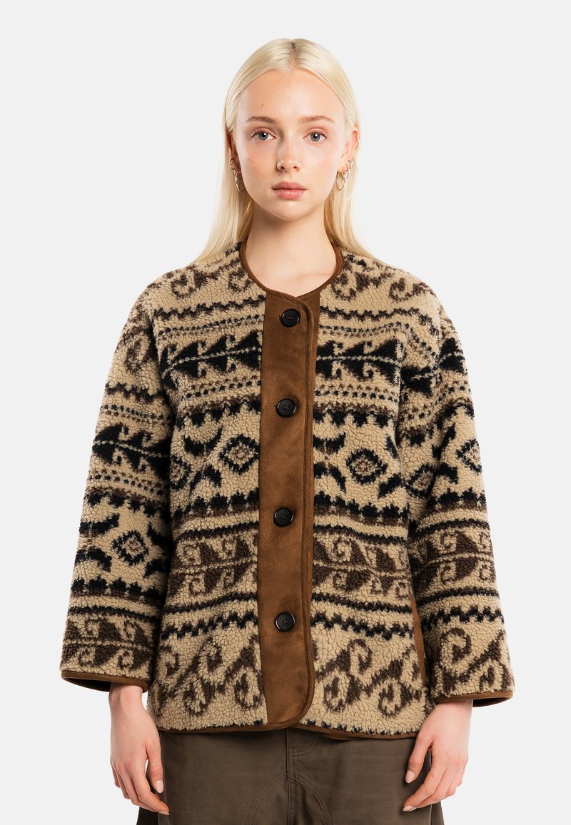 Jeune femme aux cheveux blonds raides portant une veste polaire beige et marron à boutons et un pantalon marron, debout sur un fond uni.