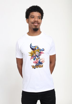 Homme portant un t-shirt blanc avec le personnage coloré Dark Magician Girl et le texte "Yu-Gi-Oh!" sur un fond blanc uni.