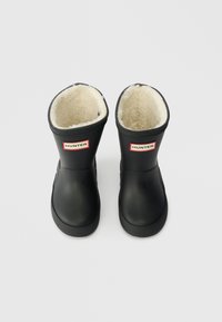 Botas de borracha pretas com um forro de pelo beje suave, apresentando um logo "HUNTER" vermelho e branco na frente. Ponta arredondada e sola texturizada.