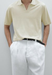 Beige polo met korte mouwen en een V-hals, gecombineerd met witte geplooide broek en een zwarte gevlochten riem. Gladde textuur, moderne pasvorm.