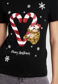 Svart t-shirt med glittrande röda och vita godiskäppar som bildar ett hjärta, en guldpaljettsloth, och texten "God Jul". Snöflingeakenter visas.