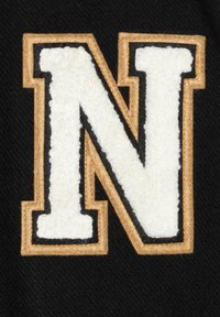 Svart tyg har en stor, upphöjd vit bokstav "N" som är kantad i guld. Den texturerade ytan ger en dimension, och de kontrasterande färgerna förbättrar synligheten.