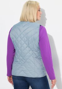 Vadderad väst i ljusblått med ett diamantmönster, bärs över en lila långärmad skjorta, i kombination med blå jeans, sedd bakifrån.