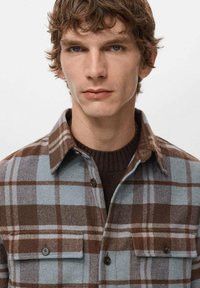 Camisa de franela a cuadros en marrón y azul claro con cuello abotonado, bolsillos frontales y textura suave sobre un suéter marrón oscuro.