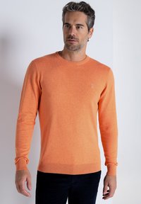 Oranger Strickpullover mit Rundhalsausschnitt, gerippten Bündchen und Saum, versehen mit einem kleinen gestickten Logo auf der Brust. Glatte Textur.