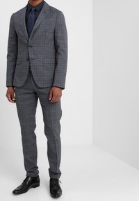Traje gris a cuadros con una chaqueta a medida y pantalones slim-fit, combinado con una camisa y corbata azul marino. Unos zapatos de cuero negros completan el look.