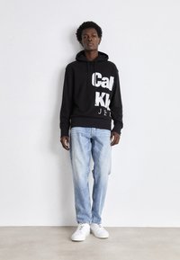 Felpa nera con testo bianco, abbinata a jeans azzurri chiari e sneakers bianche. La felpa presenta una tasca a marsupio frontale e cappuccio con lacci.