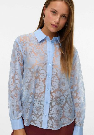 Jonge vrouw draagt een doorschijnend blauw bloesje met bloemenprint, vaste kraag en manchetten, gecombineerd met donkere rode broek.