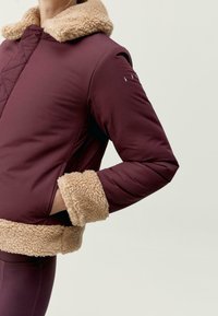 Bordový kabát s béžovým střihem vlny kolem límce a na rukávech, zdobený prošívaným designem a bočními kapsami, představující elegantní, moderní siluetu.