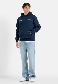 Felpa con cappuccio blu navy con tasca frontale, marchio in testo bianco. Abbinata a jeans azzurri chiari e scarpe da ginnastica bianche. Sfondo semplice.
