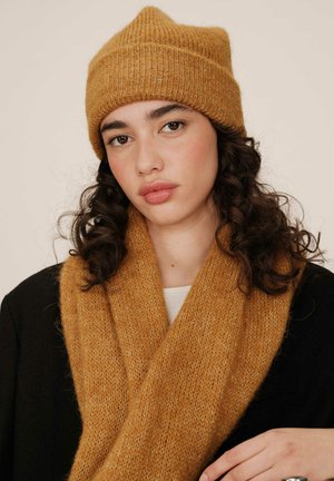 Bonnet en tricot jaune moutarde avec une texture côtelée, associé à une écharpe douce assortie, porté sur un vêtement noir. Tissu lisse et confortable.