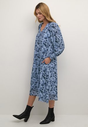 Robe à manches longues et col en V de couleur bleu clair avec des motifs floraux bleu foncé, de forme fluide, avec des poches latérales, associée à des bottines noires.