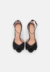 Melnas satīna augstpapēžu sandalēs ar loku priekšpusē, atvērtu purngalu un potītes siksniņu. Iespējamā gluda, matēta virsma.