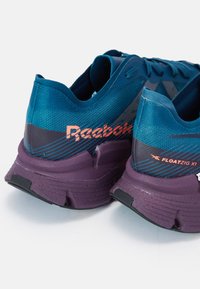 Zapatillas de correr Reebok en azul y púrpura con parte superior de malla, logo acentuado y suela texturizada. Presenta un diseño único con un talón acolchado.