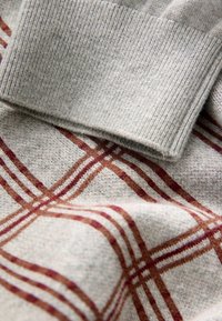 Grå strikket sweater med ribbet krave og et ternet mønster i rust og mørkerød, lavet af et blødt, tekstureret stof.
