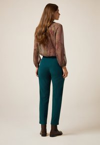 Blusa de manga larga transparente con un patrón floral apagado, combinada con pantalones ajustados en tono azul verdoso y botas de tobillo marrones; textura suave y diseño ajustado.