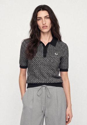 Frau mit langen, welligen dunklen Haaren, die ein kurzärmeliges Poloshirt mit schwarzem und grauem Zickzackmuster und hellgraue, gebundene Hose trägt.