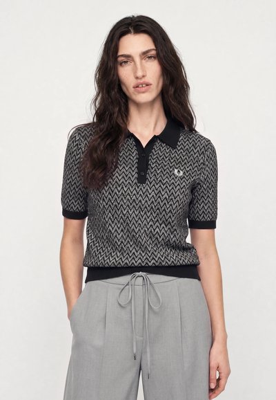 Femme aux longs cheveux ondulés et foncés portant un polo manches courtes à motif zigzag noir et gris et un pantalon gris clair noué.