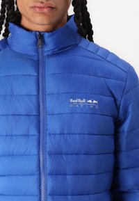 Blaues gepolstertes Jacket mit hohem Kragen, Reißverschluss und horizontalen Nähten. Verfügt über das Red Bull Racing-Logo auf der linken Brust.
