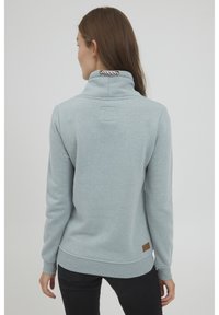 Felpa azzurra con colletto alto, polsini a coste e una patch in pelle marrone sul orlo. Tessuto morbido e con texture.