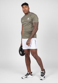 Olijfgroene getailleerde T-shirt met logo, witte sportshorts met zijbranding, houdt een zwart gewichtsschijf vast, staande op een grijze achtergrond.