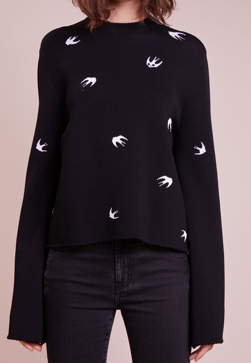Pull noir à manches longues avec des silhouettes blanches d'oiseaux associé à un jean noir sur une personne aux cheveux bruns mi-longs.