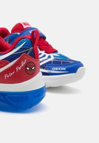 Piros és kék sportcipők hálós kialakítással, a Pókember logójával, fehér díszítésekkel és áttetsző kék talppal. Az oldalon "Peter Parker" felirat található.