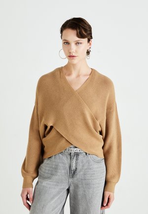 Beige omslaggenser i ribbestrikket stoff med V-hals, vide ermer og løs passform, kombinert med høytlivede grå jeans og et belte.