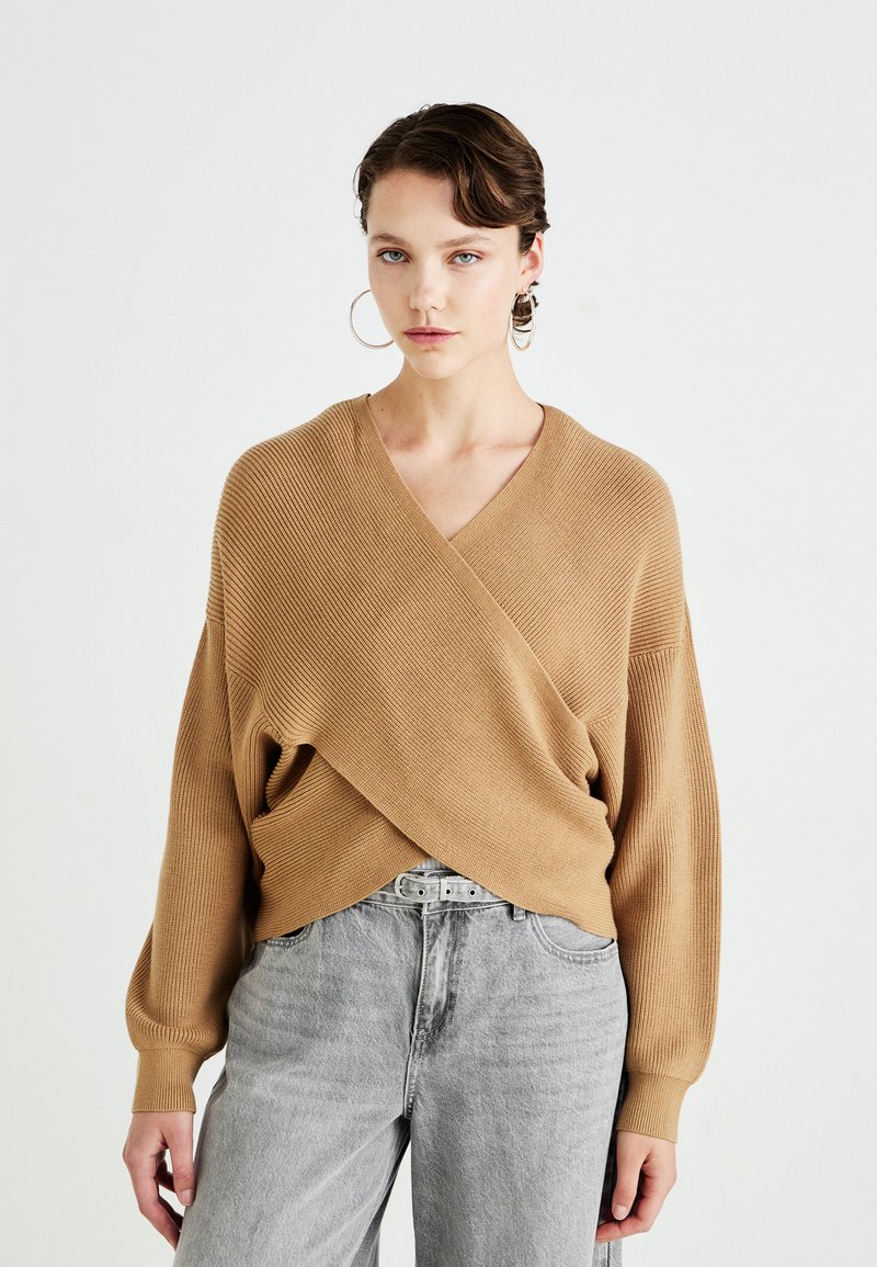 Beige omslaggenser i ribbestrikket stoff med V-hals, vide ermer og løs passform, kombinert med høytlivede grå jeans og et belte.