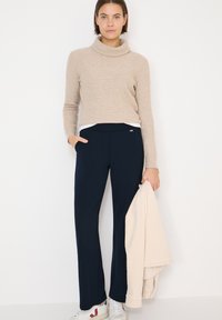 Femme portant un pull à col roulé beige, un pantalon noir, des baskets blanches, tenant une veste beige sur un fond blanc.