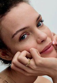 Anello in oro indossato su un dito, con pelle liscia e trucco naturale. La persona ha capelli scuri e texture e lineamenti del viso morbidi.