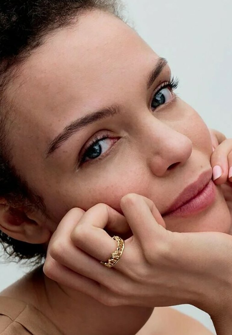 Anello in oro indossato su un dito, con pelle liscia e trucco naturale. La persona ha capelli scuri e texture e lineamenti del viso morbidi.