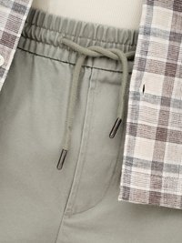 Jasnozielone spodnie z elastycznym pasem i sznurkiem oraz koszula w kremową, brązową i czarną kratę, częściowo widoczna z boku.