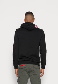 Felpa nera con cappuccio e tasca a canguro, orlo ribattuto. Presenta un'etichetta rossa sul cappuccio e dettagli grafici sulle maniche. Abbinata a pantaloni cargo color oliva.